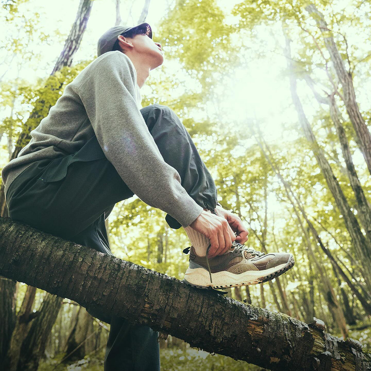 Flower MOUNTAIN (フラワーマウンテン) のイメージ画像。森の中で気に腰かけた男性がFlower MOUNTAIN (フラワーマウンテン) のスニーカーを履いている。